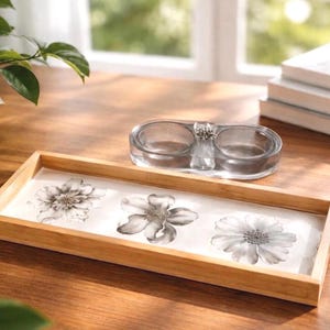 May include: A rectangular wooden tray with a light wood finish featuring three black and white floral illustrations on a white base, accompanied by a clear glass double-well dish with a small silver-toned decorative accent in the center.