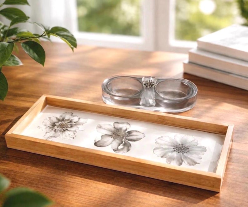 May include: A rectangular wooden tray with a light wood finish featuring three black and white floral illustrations on a white base, accompanied by a clear glass double-well dish with a small silver-toned decorative accent in the center.