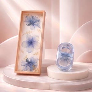 May include: A rectangular wooden tray featuring a white base with three translucent blue floral illustrations and small gold leaf accents, accompanied by a matching translucent blue ring with a floral design inside the clear resin band.