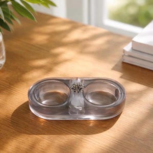 May include: A translucent, oval-shaped gray tray featuring two circular recessed compartments and a central decorative silver-toned metal accent piece with a starburst or floral design, crafted from a smooth, polished material.