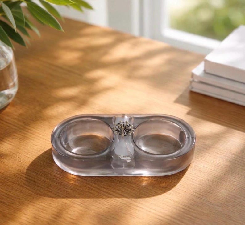 May include: A translucent, oval-shaped gray tray featuring two circular recessed compartments and a central decorative silver-toned metal accent piece with a starburst or floral design, crafted from a smooth, polished material.