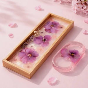 May include: A rectangular wooden tray and a small oval dish made of clear resin. Both items contain embedded pressed purple pansies, small white flowers, and sprigs of lavender. The items are set against a light pink background with scattered petals.