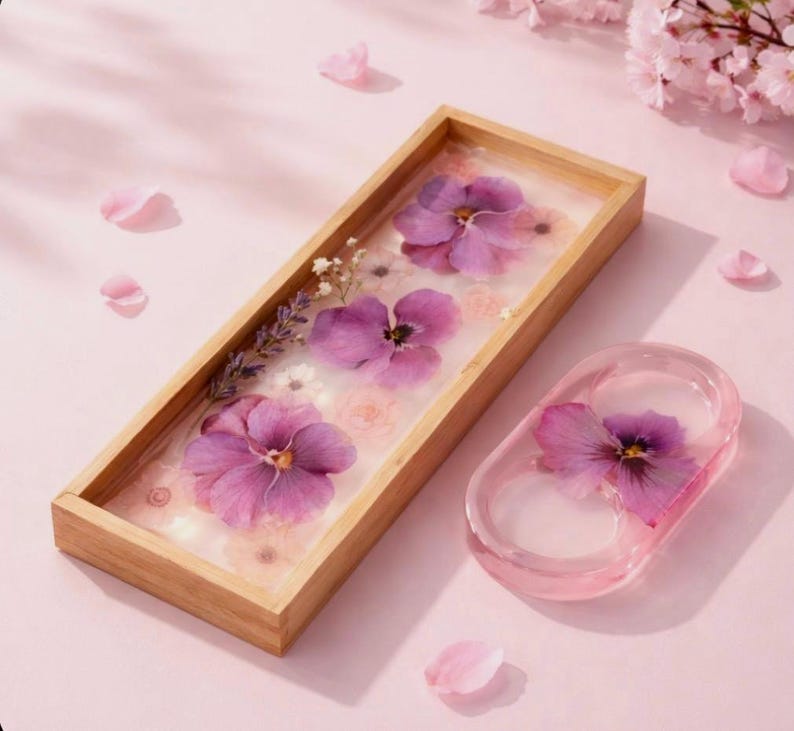 May include: A rectangular wooden tray and a small oval dish made of clear resin. Both items contain embedded pressed purple pansies, small white flowers, and sprigs of lavender. The items are set against a light pink background with scattered petals.