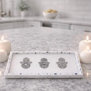 May include: A rectangular white tray with a speckled blue and white pattern, featuring three black Hamsa hand designs. The tray is surrounded by three lit white candles, creating a serene and decorative display.