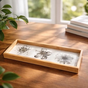 May include: A rectangular wooden tray with a light brown frame. The tray's interior features a white surface with three grayscale floral designs. The flowers are in full bloom, with detailed petals and centers.