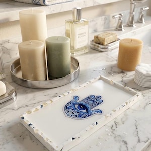 May include: A white rectangular tray with a blue and white Hamsa hand design. The tray is surrounded by a speckled border. Several pillar candles in shades of beige and green sit on a silver tray. A bottle of bath oil and soap are also visible.