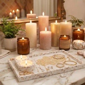 May include: A collection of lit candles in various sizes and colors, including pillar candles and jar candles, arranged on a marble countertop. A decorative tray holds jewelry. A diffuser and a small potted plant are also present.