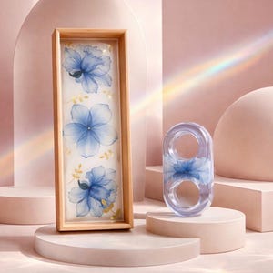 May include: A rectangular wooden tray featuring a white base with three blue floral illustrations and small gold leaf accents, paired with a clear, rounded resin object containing a matching blue floral design inside its structure.