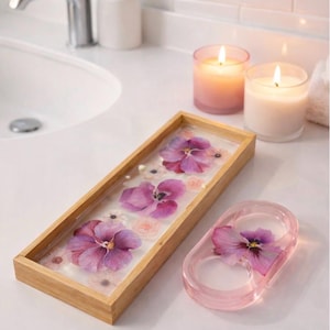 May include: A rectangular wooden tray with a clear resin base containing pressed purple pansies and small white flowers, alongside a matching oval-shaped pink resin ring holder featuring a single pressed purple pansy embedded inside.