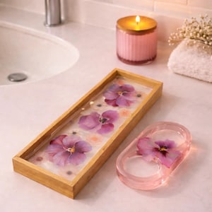 May include: A rectangular wooden tray with a clear resin base containing pressed purple pansies and small white flowers, alongside a matching oval-shaped pink resin ring holder featuring a single embedded purple pansy flower.