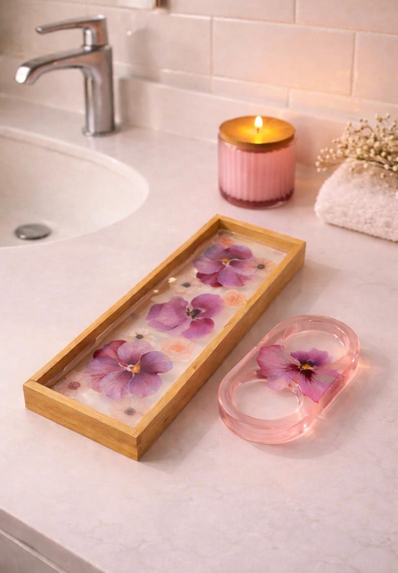 May include: A rectangular wooden tray with a clear resin base containing pressed purple pansies and small white flowers, alongside a matching oval-shaped pink resin ring holder featuring a single embedded purple pansy flower.