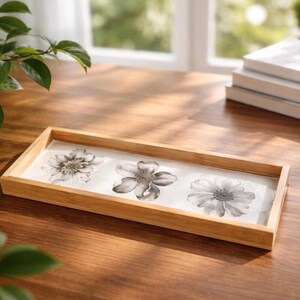 May include: A rectangular wooden tray with a light brown finish and a white base. The interior of the tray features a decorative print of three distinct black and white floral illustrations arranged in a horizontal line across the center.