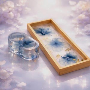 May include: A rectangular wooden tray and a matching oval resin candle holder, both featuring embedded blue floral patterns and delicate gold leaf accents. The tray has a light wood frame, while the candle holder is clear, rounded resin.