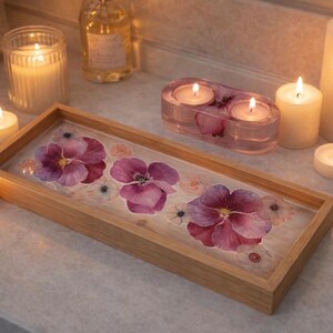 May include: A rectangular wooden tray featuring a clear resin base embedded with pressed purple and pink pansy flowers. The tray is surrounded by several lit white pillar and tea light candles, along with a small oval resin candle holder.