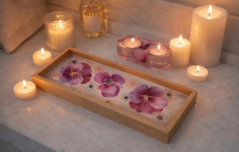 May include: A rectangular wooden tray featuring a clear resin base embedded with pressed purple and pink pansy flowers. The tray is surrounded by several lit white pillar and tea light candles, along with a small oval resin candle holder.