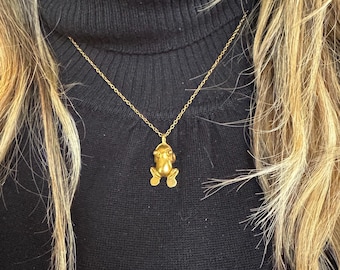 May include: A gold-colored frog pendant necklace. The frog charm is detailed with visible eyes and legs, suspended from a delicate gold chain. The necklace is worn against a black turtleneck.