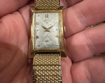 Puede incluir: Un reloj de pulsera Lord Elgin vintage en tono dorado. La esfera rectangular tiene una esfera plateada con manecillas e indicadores de hora dorados. La correa es de metal tejido en tono dorado. La esfera del reloj dice "Lord Elgin".