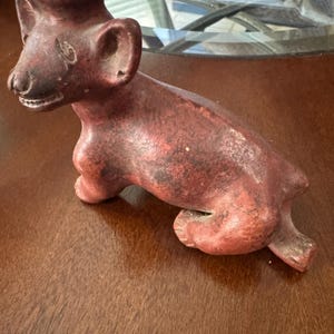 Op de afbeelding: Een roodbruin terracotta dierenbeeldje in de vorm van een hond of coyote, met een holle cilindrische opening op de kop, een getextureerd mat oppervlak, puntige oren en een zittende houding met de staart zijwaarts uitgestrekt.