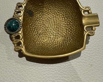 Puede incluir: Un cenicero de latón con una textura martillada en la superficie interior. La pieza presenta volutas decorativas en los lados y una incrustación de piedra circular de color verde oscuro moteado en un extremo, con una muesca en el lado opuesto.