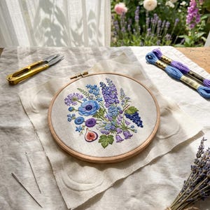 Puede incluir: Un bastidor de bordado redondo de madera con tela color hueso que presenta un diseño floral detallado con flores azules y moradas, hojas verdes y un higo cortado. Se incluyen tijeras doradas, dos agujas y dos madejas de hilo de bordar.