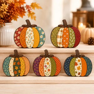 May include: Five flat, decorative pumpkin-shaped ornaments featuring various fabric patterns in autumn colours including orange, yellow, green, and red. Each pumpkin has a dark brown wooden stem and is divided into vertical segments with floral, leaf, and plaid prints.