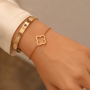 Puede incluir: Una pulsera rígida de metal en tono dorado con recortes circulares y piedras transparentes alternadas, junto a una pulsera de cadena fina en tono dorado con un motivo de trébol de cuatro hojas en el centro. Ambas piezas tienen acabado pulido.