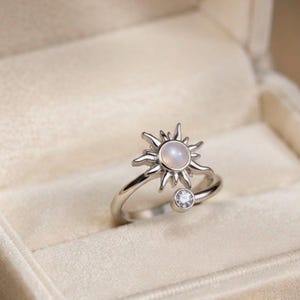 Puede incluir: Un anillo abierto de tono plateado con un motivo en forma de sol que presenta una piedra central redonda, lisa y pálida. El extremo opuesto de la banda está acentuado con una pequeña piedra preciosa redonda, clara y facetada.