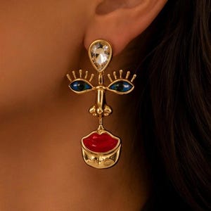 Puede incluir: Un pendiente dorado con forma de rostro estilizado. La parte superior presenta un cristal transparente en forma de lágrima, seguido de dos ojos azules almendrados con pestañas doradas, una nariz central y labios rojos brillantes.