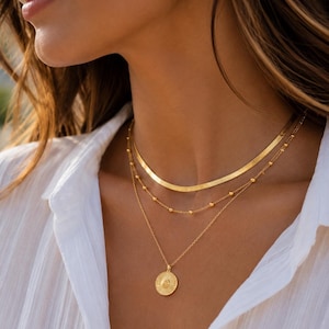 Op de afbeelding: Een gelaagde gouden kettingenset bestaande uit drie afzonderlijke kettingen. De bovenste laag is een platte visgraatketting, de middelste laag is een fijne ketting met kleine, gelijkmatig verdeelde kraaltjes en een ronde hanger met zonnemotief.