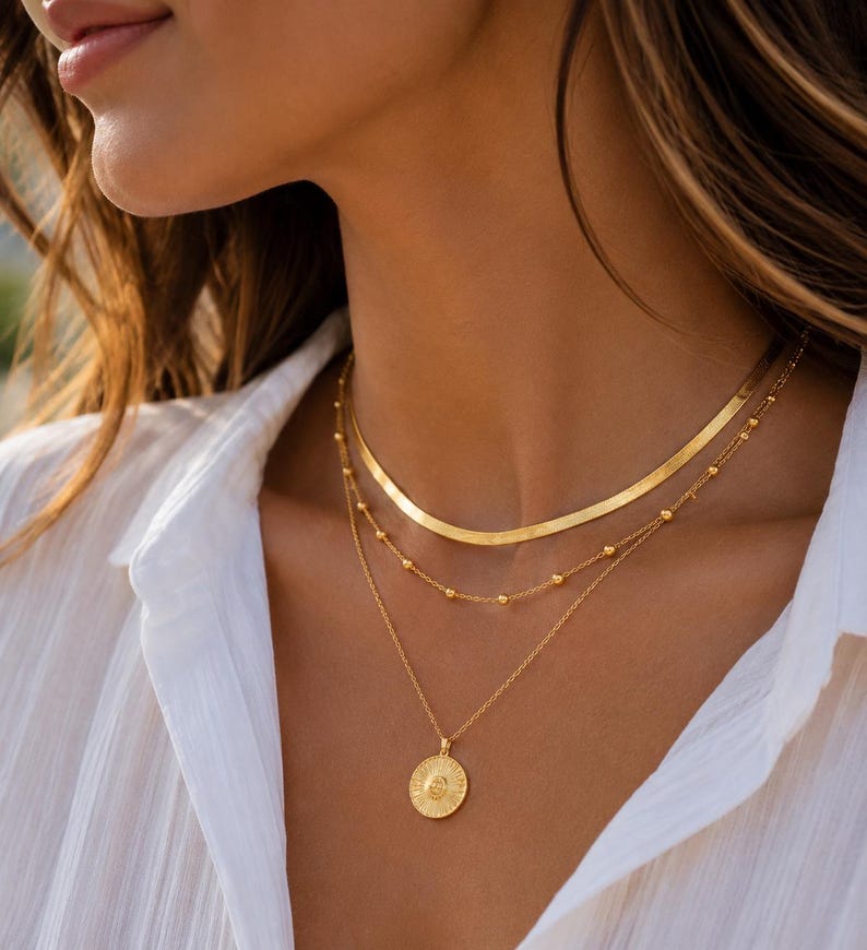 Op de afbeelding: Een gelaagde gouden kettingenset bestaande uit drie afzonderlijke kettingen. De bovenste laag is een platte visgraatketting, de middelste laag is een fijne ketting met kleine, gelijkmatig verdeelde kraaltjes en een ronde hanger met zonnemotief.