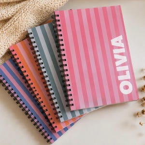 Puede incluir: Una pila de cuatro cuadernos de espiral con rayas verticales en rosa, azul grisáceo, naranja y morado. El cuaderno superior tiene el nombre OLIVIA impreso en letras mayúsculas blancas grandes en el lado derecho de la cubierta a rayas rosas.
