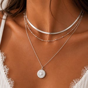 Op de afbeelding: Een gelaagde zilveren kettingenset bestaande uit drie verschillende kettingen. De bovenste laag is een platte visgraatketting, de middelste is een fijne kralenketting en de onderste is een fijne schakelketting met een ronde zonhanger.