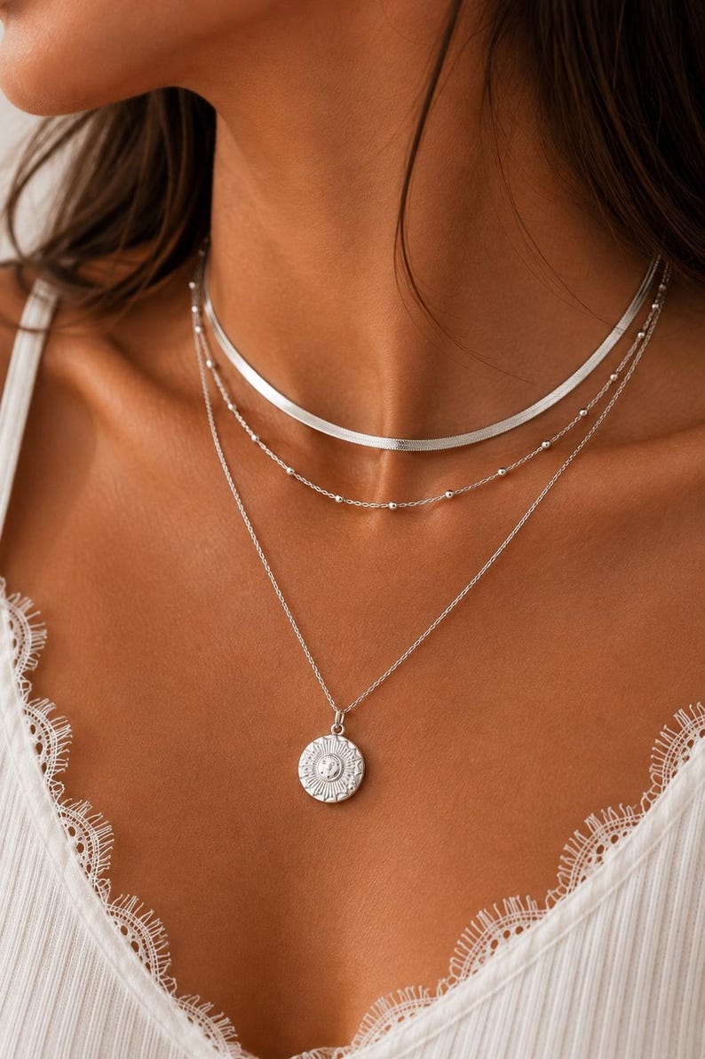 Op de afbeelding: Een gelaagde zilveren kettingenset bestaande uit drie verschillende kettingen. De bovenste laag is een platte visgraatketting, de middelste is een fijne kralenketting en de onderste is een fijne schakelketting met een ronde zonhanger.