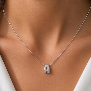 Puede incluir: Un collar de cadena en tono plateado con un pequeño colgante redondeado en forma de letra mayúscula A. El colgante está incrustado con numerosas piedras diminutas, claras y brillantes que cubren toda su superficie frontal, creando un aspecto luminoso.