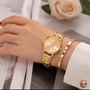 Puede incluir: Un reloj analógico de tono dorado con correa de eslabones a juego y esfera redonda con marcadores finos, acompañado de una pulsera rígida y delgada de tono dorado grabada con las letras L, O, O y E en secuencia vertical sobre la banda.
