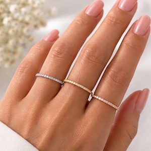 Puede incluir: Tres anillos finos y delicados en tres dedos. Un anillo es de tono plateado, otro de tono dorado y otro de tono oro rosa. Cada anillo presenta una hilera continua de pequeñas piedras claras y brillantes engastadas a lo largo de la banda.