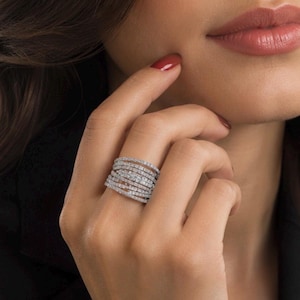 Puede incluir: Un anillo ancho de múltiples bandas hecho de metal plateado, con numerosas bandas finas y superpuestas incrustadas con pequeñas piedras claras y brillantes dispuestas en un diseño continuo, estratificado y texturizado en el dedo.