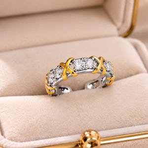 Puede incluir: Un anillo abierto de tono plateado con piedras redondas transparentes alternadas con detalles metálicos dorados en forma de X. El anillo tiene un acabado pulido y una abertura en la parte inferior, mostrando una serie de piedras brillantes.