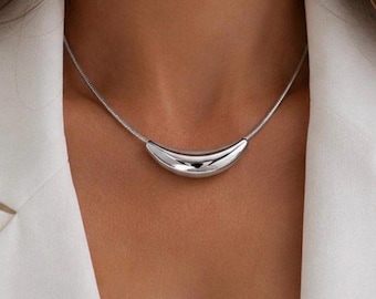 Op de afbeelding: Een zilverkleurige ketting met een gladde, gebogen, halvemaanvormige hanger aan een dunne, flexibele ketting. Het metalen sieraad heeft een gepolijst, reflecterend oppervlak dat het licht opvangt bij het sleutelbeen.