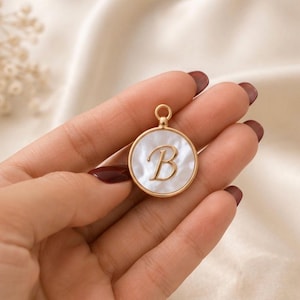Puede incluir: Un colgante circular con una incrustación de nácar blanco engastada en un marco de metal dorado. El centro del colgante muestra una letra B mayúscula en relieve de color dorado en fuente cursiva, con una pequeña anilla circular en la parte superior.
