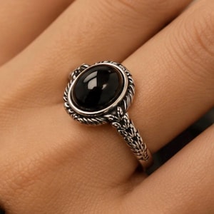 Puede incluir: Un anillo de plata con una piedra negra lisa de forma ovalada en el centro. La banda de metal y el engaste que rodea la piedra están decorados con intrincados patrones texturizados que recuerdan a una cuerda trenzada y grabados de hojas.