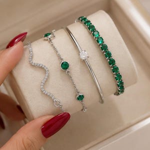 Puede incluir: Un conjunto de cuatro pulseras de tono plateado. Una presenta una línea ondulada de piedras claras, otra es una cadena delicada con dos piedras verdes redondas, la tercera es un brazalete liso con una piedra clara y la cuarta es una pulsera de tenis con piedras verdes redondas.