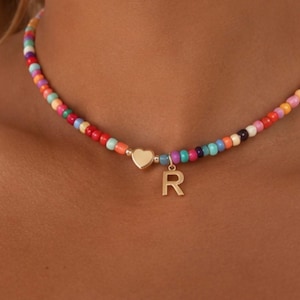 Puede incluir: Un collar tipo gargantilla con una hilera de pequeñas cuentas de vidrio multicolores en tonos rojo, naranja, amarillo, verde, azul y morado. El collar incluye un dije de corazón dorado y un colgante con la letra R en tono dorado.
