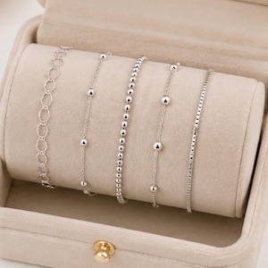 Puede incluir: Un conjunto de cinco pulseras de tono plateado exhibidas en un rollo de joyería de terciopelo beige. La colección incluye una cadena con eslabones circulares abiertos, dos cadenas finas con pequeñas cuentas esféricas, una cadena de cuentas y una cadena cuadrada.