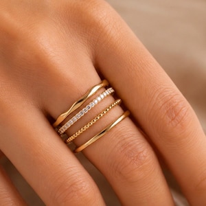 Puede incluir: Un conjunto de cuatro anillos dorados apilados en un dedo. El conjunto incluye una banda ondulada lisa, una banda incrustada con pequeñas piedras transparentes, una banda con textura de cuentas y una banda de oro pulido simple y brillante.