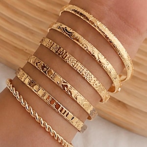 Puede incluir: Un conjunto de seis brazaletes de metal en tono dorado que se llevan en la muñeca. La colección incluye cinco pulseras finas con varios patrones y texturas grabadas, y una pulsera con un diseño de cuerda trenzada. Cada pieza tiene un acabado pulido.