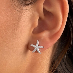 Puede incluir: Un pendiente de botón con forma de estrella de mar plateada que presenta una superficie texturizada con pequeños acentos reflectantes. El diseño de cinco puntas se sitúa en el lóbulo, mostrando un acabado metálico que capta la luz.