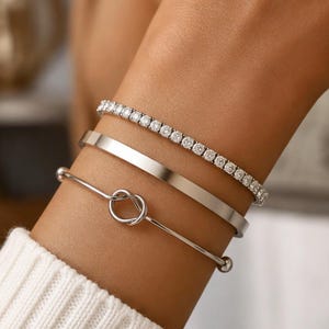 Puede incluir: Un conjunto de tres pulseras plateadas usadas juntas. La pulsera superior es de estilo tenis con una hilera de piedras redondas transparentes. La pulsera central es un brazalete de metal liso y sólido. La inferior es una pulsera fina con un nudo central.