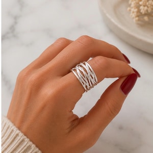 Puede incluir: Un anillo de plata con un diseño tejido. El anillo presenta múltiples bandas de plata que se entrelazan para crear un patrón calado y texturizado. El anillo se lleva en un dedo con esmalte de uñas rojo.