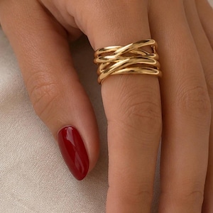 Puede incluir: Un anillo dorado con un diseño trenzado se muestra en una mano con esmalte de uñas rojo. El anillo tiene múltiples bandas entrelazadas, creando un aspecto texturizado y elegante. El anillo es una joya.